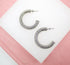 A pair of textured silver hoop earrings rests on a white display surface against a soft pink background.
