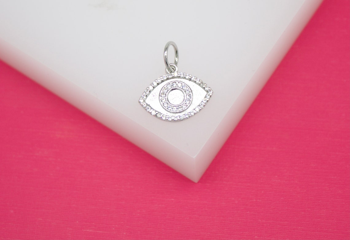 A sparkling silver evil eye charm rests on a white display against a vibrant pink background.