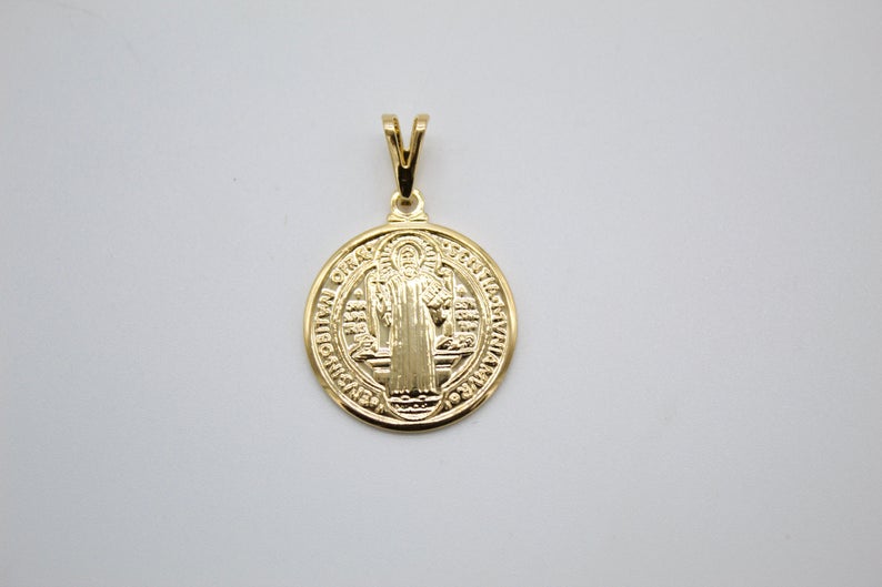 A polished gold Saint Benedict medal pendant displays intricate religious engravings against a neutral background.