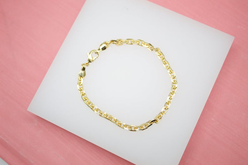 A polished gold link bracelet rests neatly on a white display pad against a soft pink background.