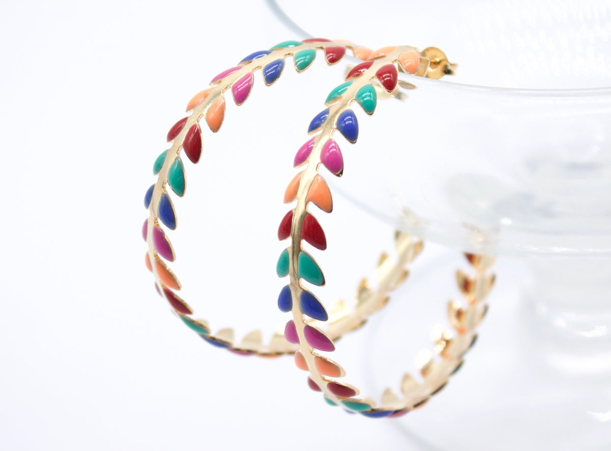 A pair of gold hoop earrings features vibrant, multicolored enamel leaf details against a bright white background.