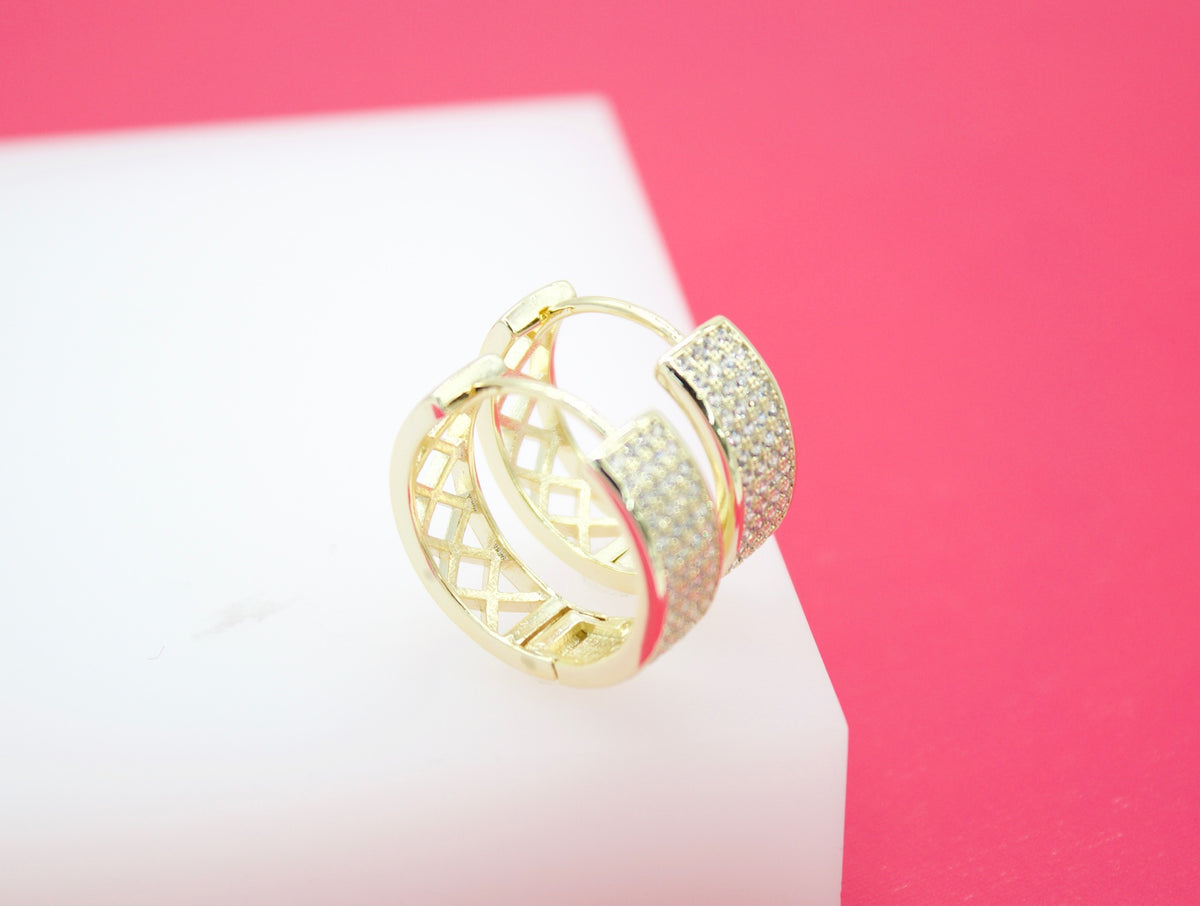 A pair of gold huggie hoop earrings featuring pavé stones and a geometric lattice pattern rests on a white display block against a vibrant pink background.