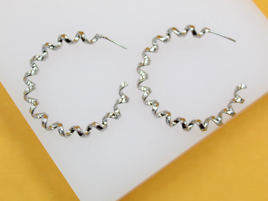 A pair of glossy silver spiral hoop earrings rests on a white display surface against a warm yellow background.