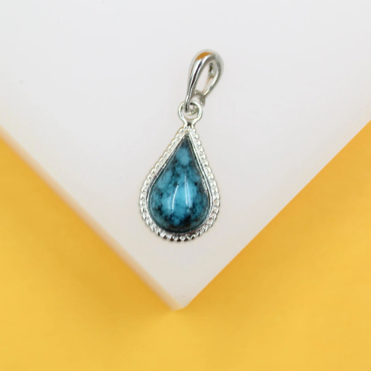 A teardrop-shaped turquoise gemstone pendant with a silver bezel rests against a clean, neutral background.