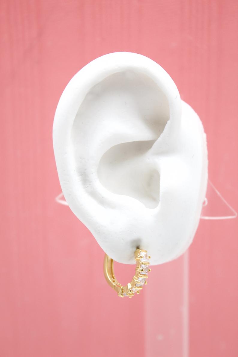 A white mannequin ear displays a gold huggie hoop earring adorned with sparkling cubic zirconia stones against a pink background.