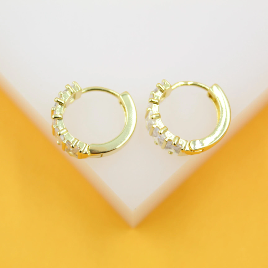 A pair of gold huggie hoop earrings embedded with sparkling cubic zirconia stones rests neatly against a white and orange display stand.