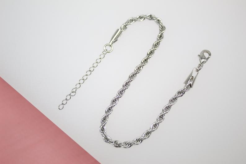 A shiny silver rope chain anklet with an adjustable extension link rests against a neutral white and pink surface.