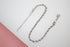 A shiny silver rope chain anklet with an adjustable extension link rests against a neutral white and pink surface.