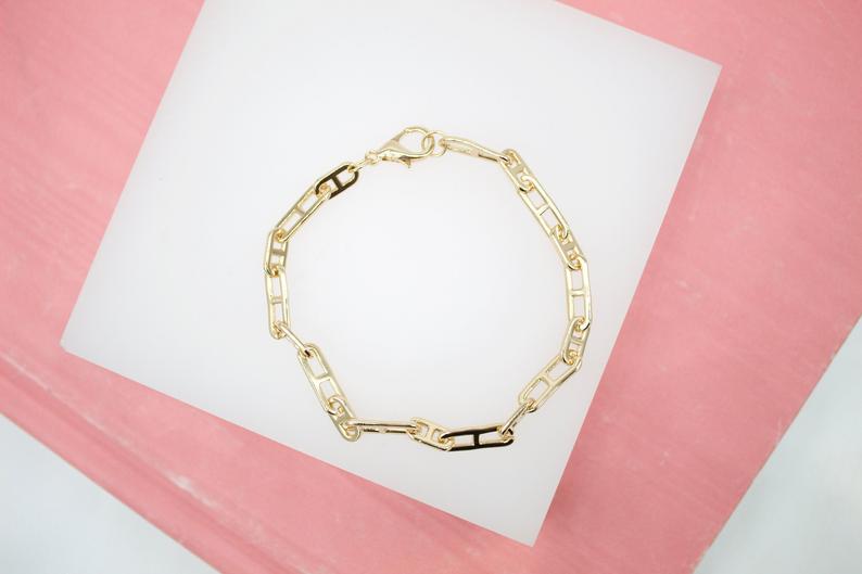 A gold link bracelet with H-shaped motifs rests on a white display surface against a pink background.