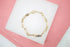 A gold link bracelet with H-shaped motifs rests on a white display surface against a pink background.