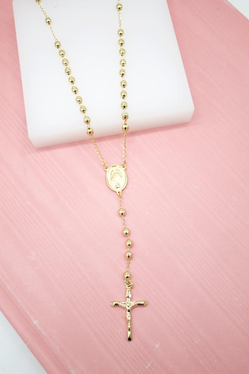 A gold-plated Catholic rosary necklace featuring faceted bead decades and a crucifix rests on a white display block against a pink background.
