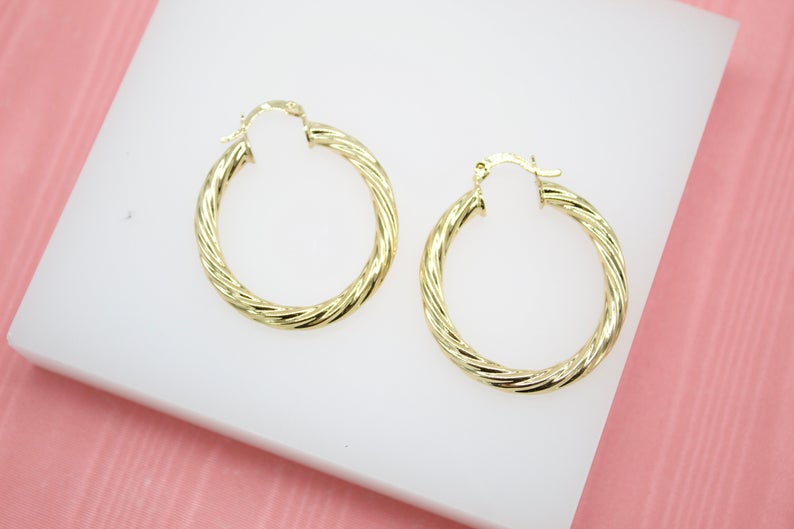 A pair of polished gold twist hoop earrings rests on a white display against a pink background.