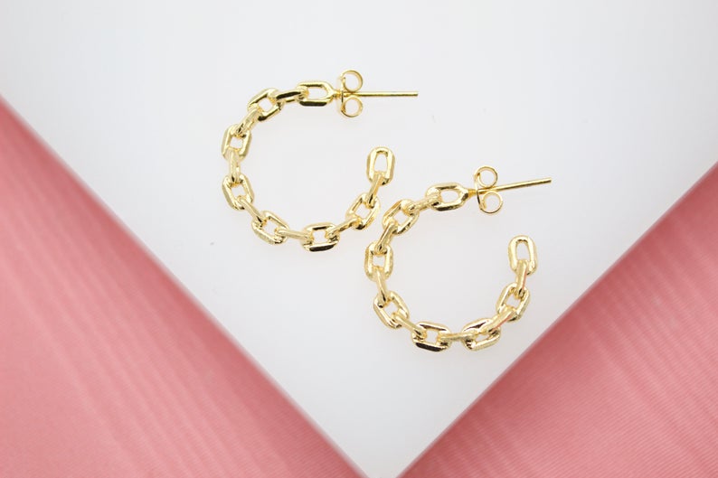 A pair of gold chain-link hoop earrings rests on a white and pink display stand.