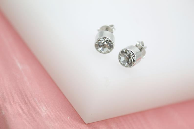 A pair of sparkling silver stud earrings rests on a white display block against a soft pink backdrop.