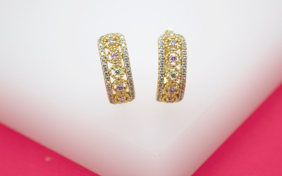 A pair of ornate gold hoop earrings features intricate filigree work, sparkling clear cubic zirconia borders, and delicate pink and green gemstones centered in floral motifs.