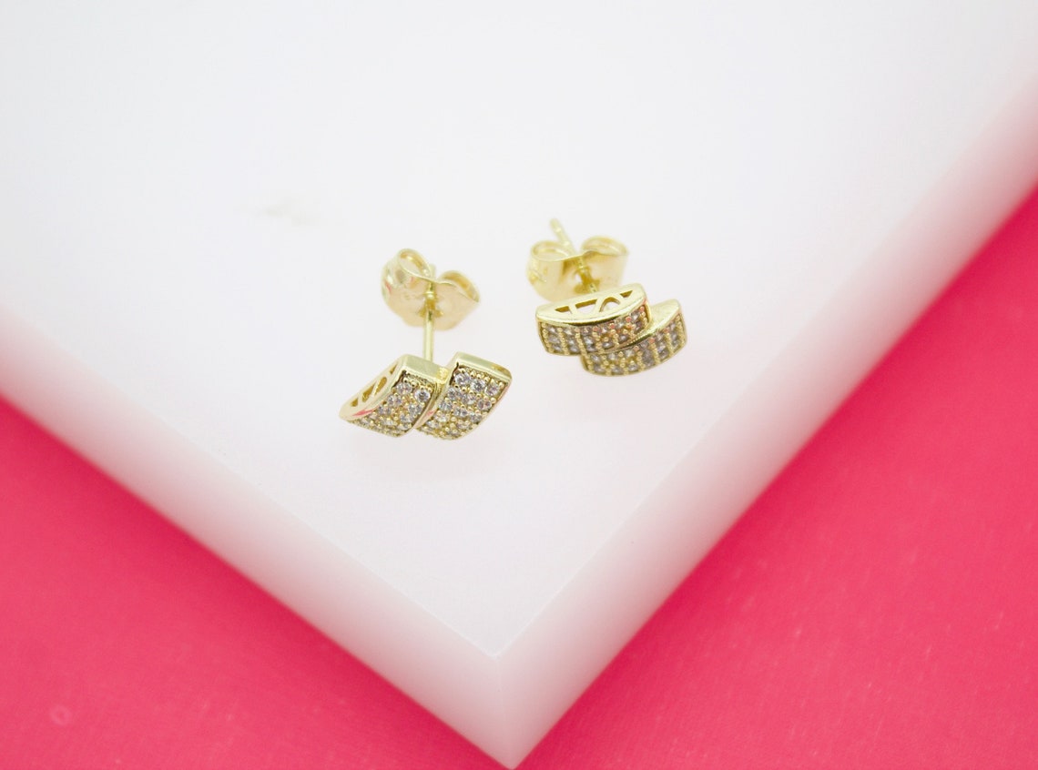 A pair of gold stud earrings featuring curved, pave-set cubic zirconia bars resting on a white display surface against a pink background.