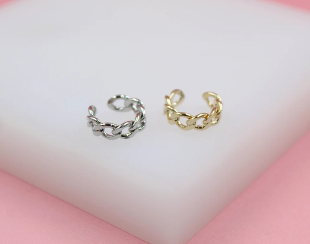 A silver and a gold chain-link ear cuff rest side-by-side on a white display surface against a pink background.