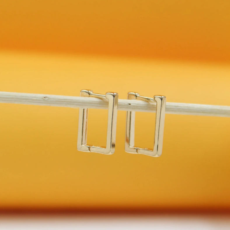 Two sleek gold rectangular hoop earrings hang side-by-side on a thin white stick against a warm orange background.