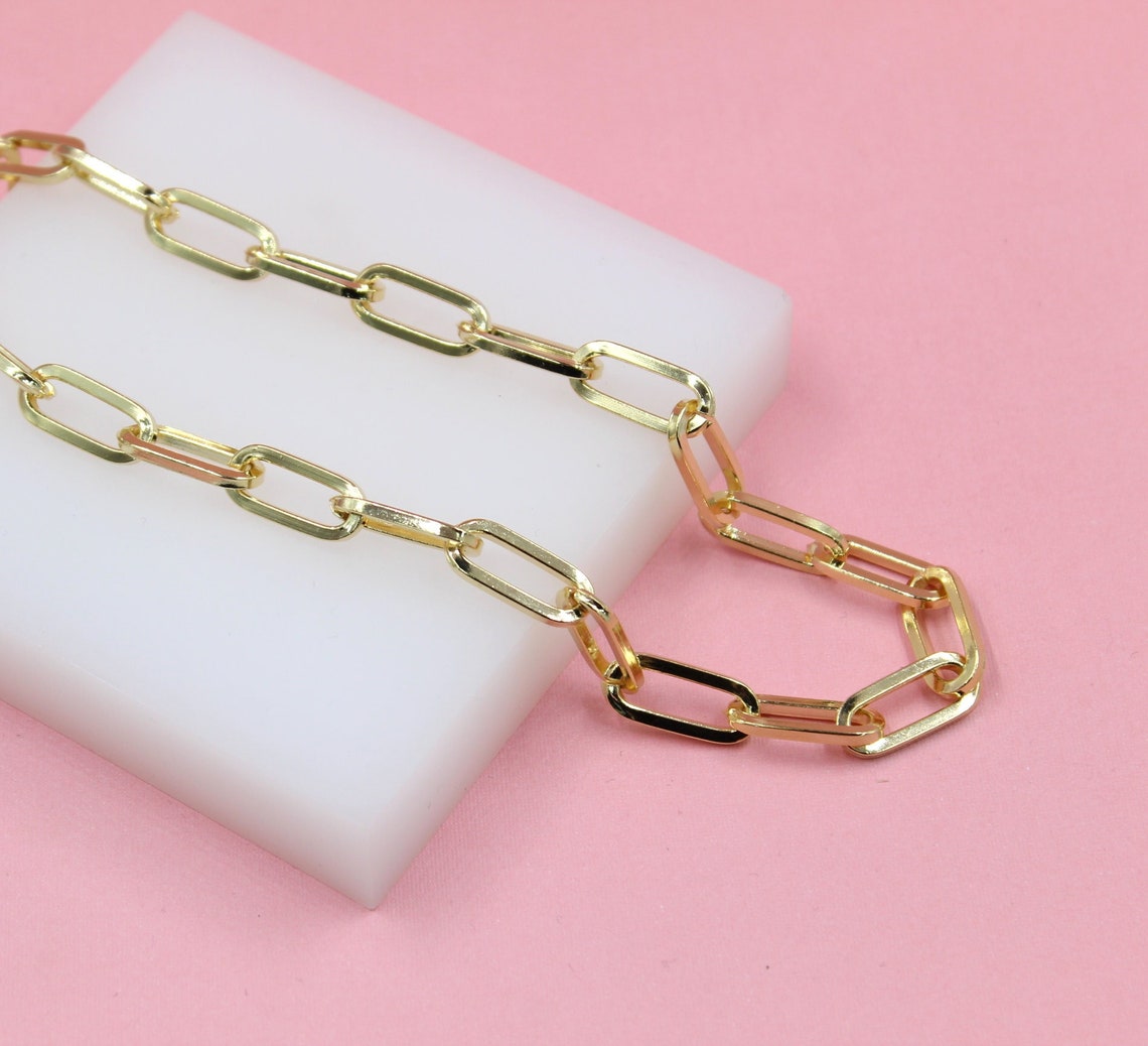 A glossy gold paperclip chain necklace rests on a white block against a soft pink background.