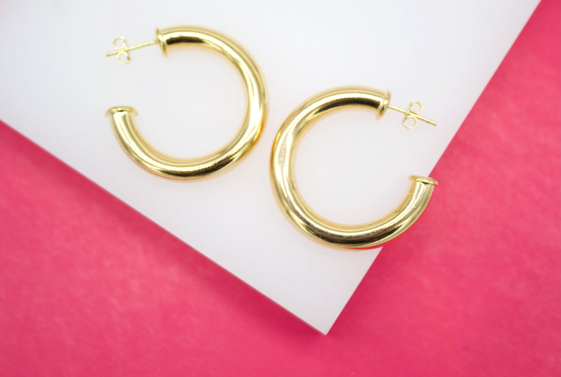 A pair of shiny gold hoop earrings rests on a white display block against a vibrant pink background.