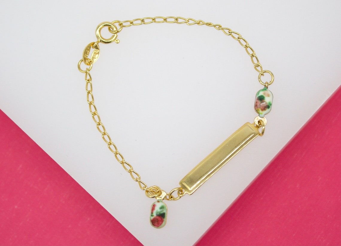 A delicate gold ID bracelet rests on a white and pink display surface, featuring an engraved rectangular plate and floral enamel bead accents.