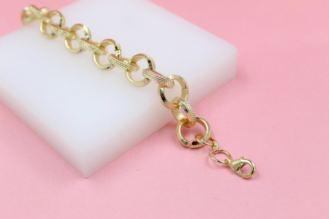 A gold-plated chain link bracelet rests on a white display block against a soft pink background.