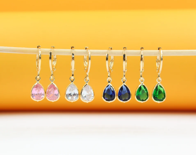 Four pairs of gold huggie hoop earrings featuring colorful teardrop-shaped cubic zirconia stones hang in a row against a vibrant yellow and white background.