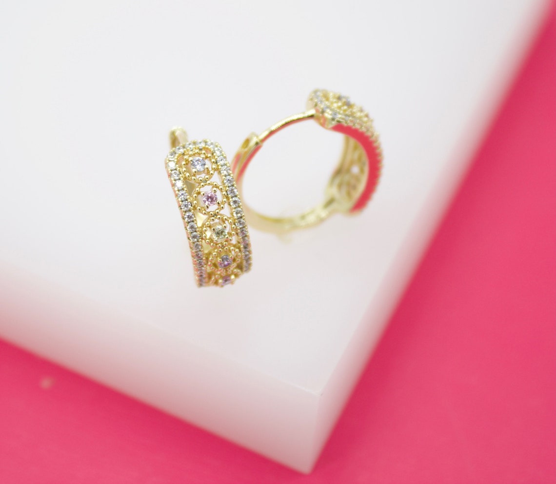 A pair of gold huggie hoop earrings featuring intricate filigree work, sparkling pavé stones, and colorful accents rests on a white display against a pink background.