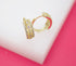 A pair of gold huggie hoop earrings featuring intricate filigree work, sparkling pavé stones, and colorful accents rests on a white display against a pink background.