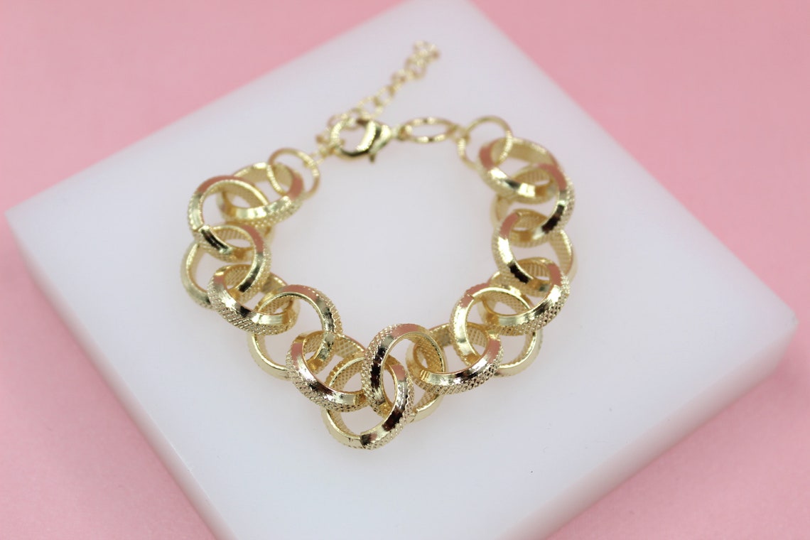 A gold-plated chunky link bracelet rests on a white display block against a soft pink background.