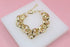 A gold-plated chunky link bracelet rests on a white display block against a soft pink background.