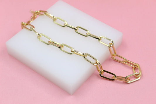 A shiny gold paperclip chain necklace rests on a white display block against a soft pink background.