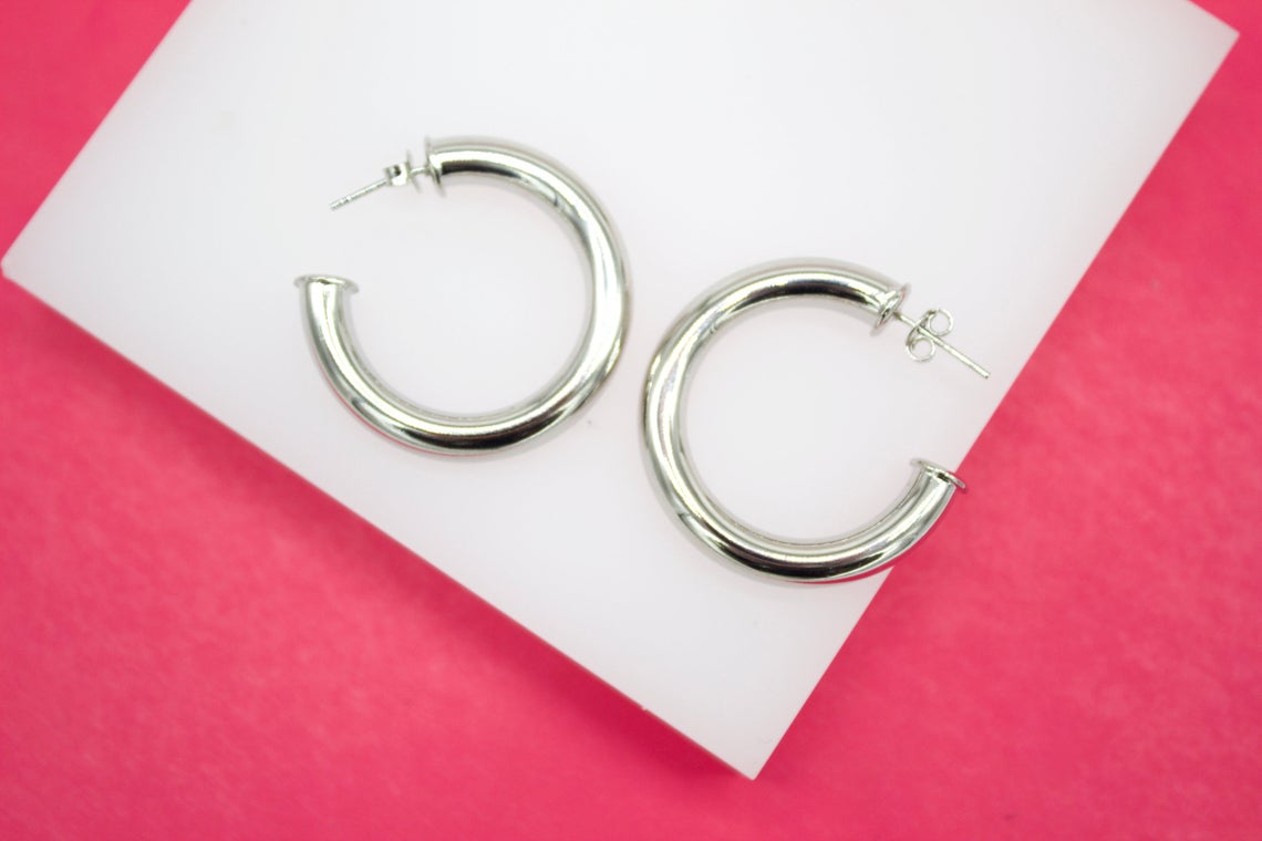 A pair of polished silver chunky hoop earrings rests on a white display against a vibrant pink background.