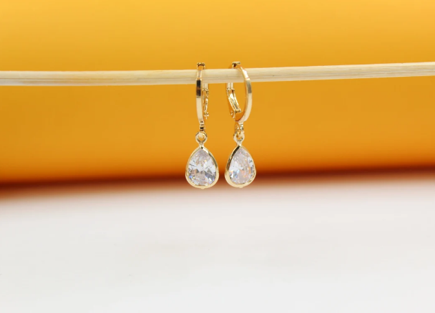 A pair of gold teardrop earrings with sparkling cubic zirconia stones hangs delicately from a wooden stick against a warm yellow and white background.
