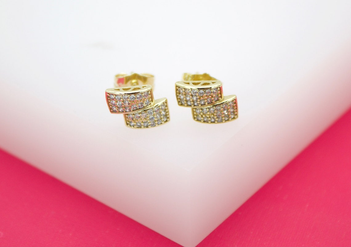 A pair of gold stud earrings featuring a chic double-bar design encrusted with sparkling cubic zirconia stones rests on a white display surface.