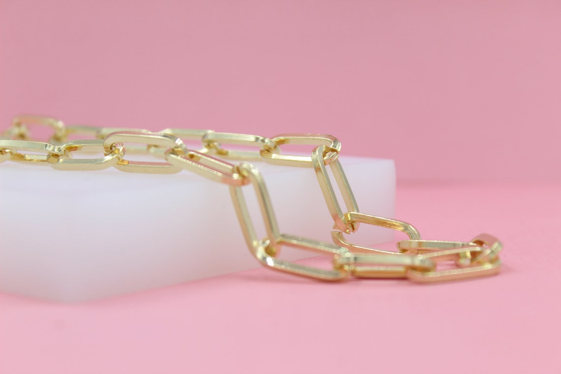 A shiny gold paperclip chain necklace rests elegantly on a white display block against a soft pink background.
