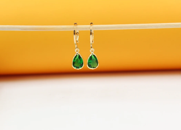 A pair of gold-plated huggie hoop earrings featuring dangling teardrop-shaped green gemstone pendants hangs delicately from a light-colored wooden stick against a warm orange backdrop.