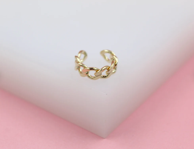 A gold chain link ear cuff rests on a white display block against a soft pink background.