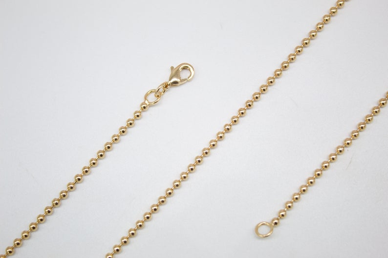 A gold-colored ball chain necklace rests against a neutral white background, showcasing its clasp and delicate spherical links.