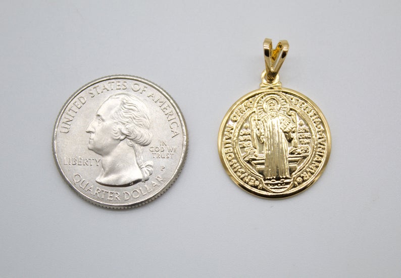A gold Saint Benedict pendant rests beside a US quarter coin to show its size.
