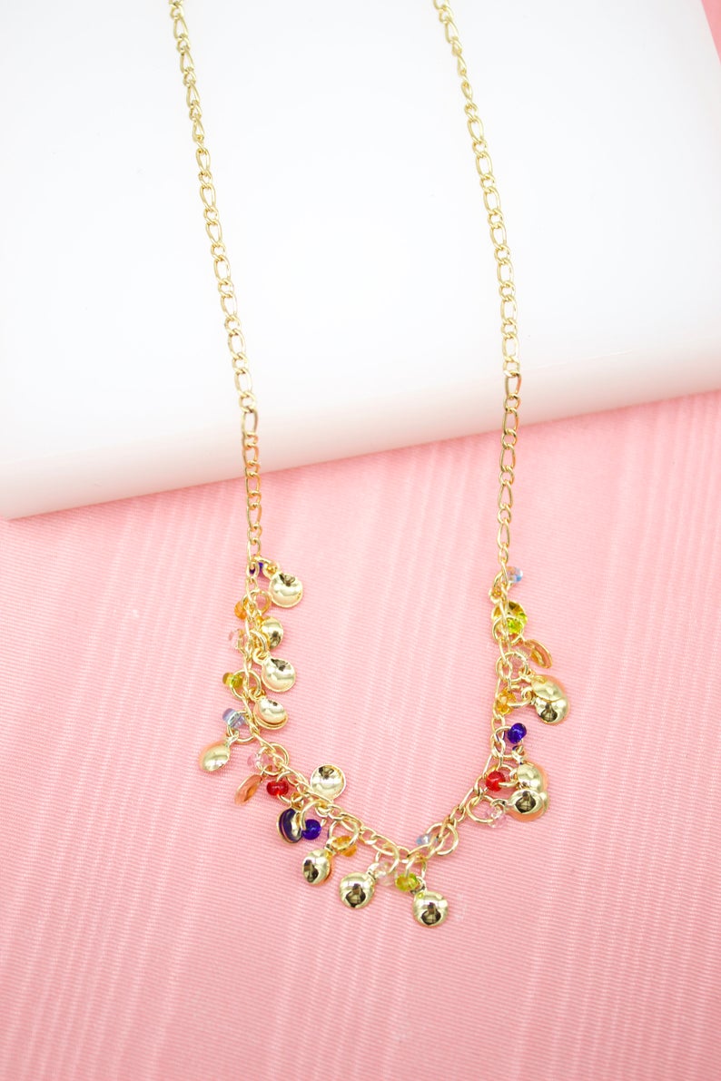 A delicate gold charm necklace featuring dangling metallic discs and colorful beads rests against a pink background.