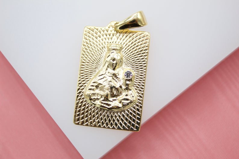 A rectangular gold pendant features a textured engraving of the Virgin Mary holding baby Jesus with a small crystal accent.