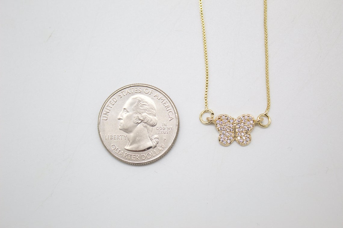 A delicate gold chain necklace with a diamond-pave butterfly pendant rests next to a US quarter for size comparison.