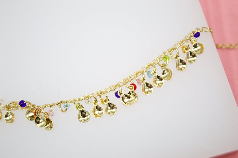 A delicate gold charm anklet features dangling golden discs interspersed with tiny multi-colored beads against a white and pink background.