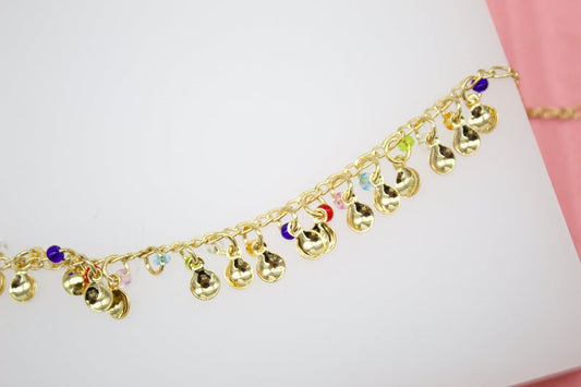 A delicate gold charm anklet features dangling golden discs interspersed with tiny multi-colored beads against a white and pink background.