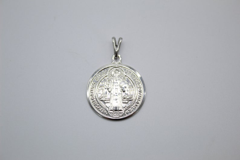 A shiny silver Saint Benedict medal pendant rests against a plain grey background, showcasing intricate religious engraving on its circular surface.