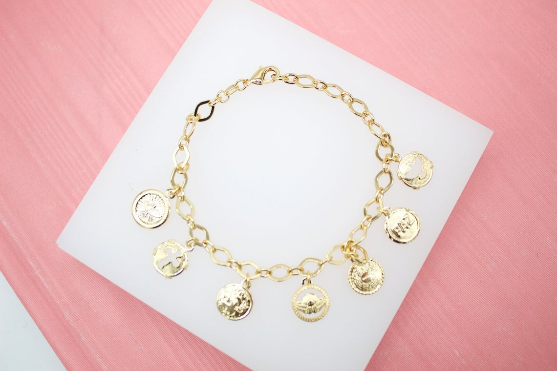 A gold charm bracelet with religious medals lies on a white display block against a pink background.