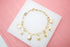 A gold charm bracelet with religious medals lies on a white display block against a pink background.
