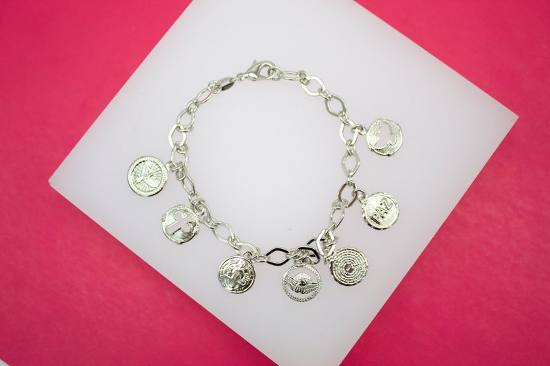 A silver charm bracelet featuring religious and inspirational medals rests on a white display against a vibrant pink background.