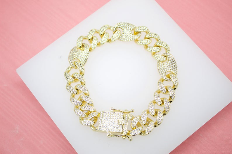 A gold-plated iced out cuban link bracelet rests on a white display surface against a pink background.